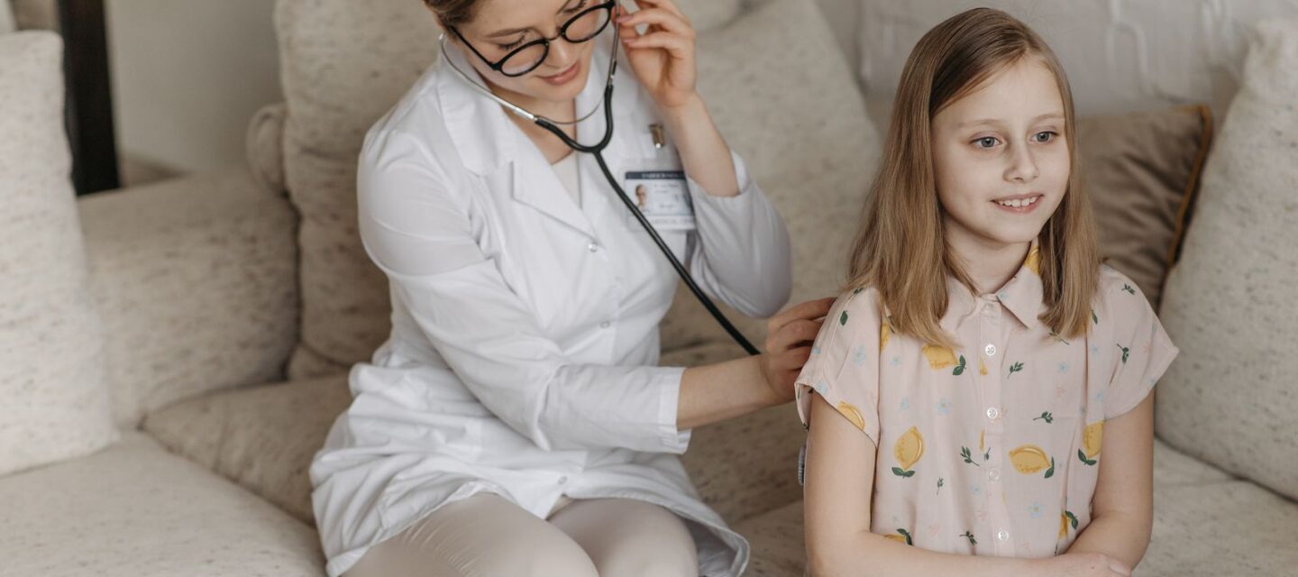 Doctor in a white coat using a stethoscope to listen to a young girl's lungs during a calm home or clinic examination