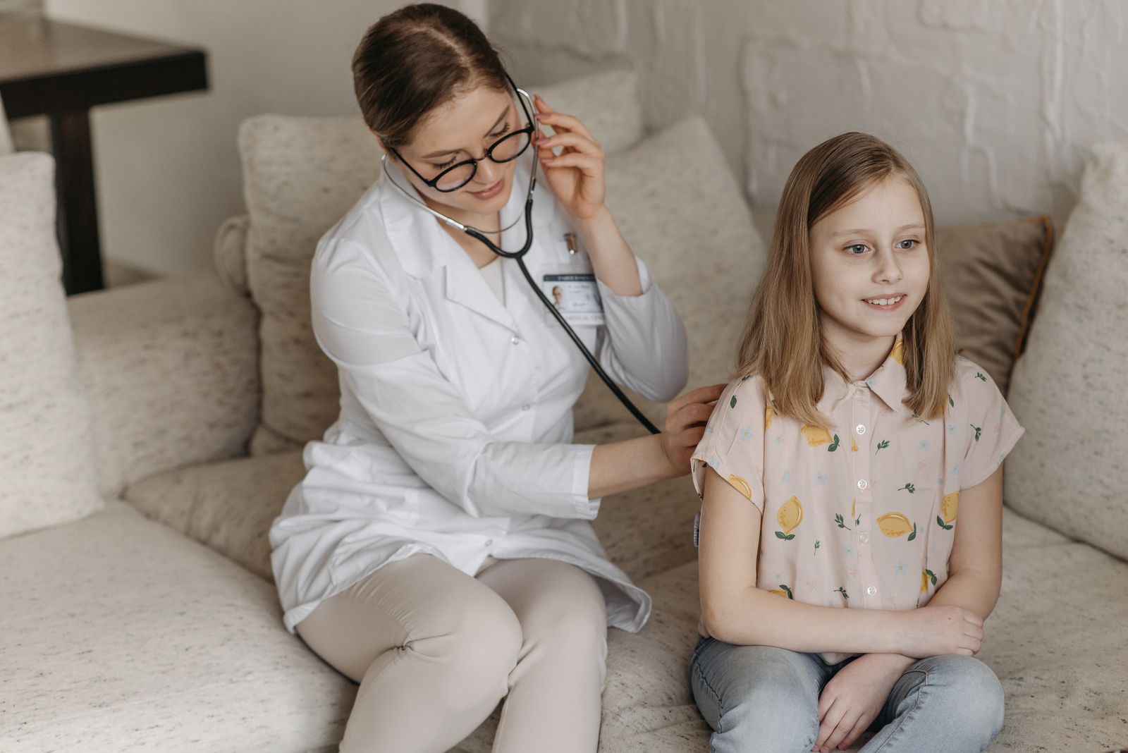 Doctor in a white coat using a stethoscope to listen to a young girl's lungs during a calm home or clinic examination