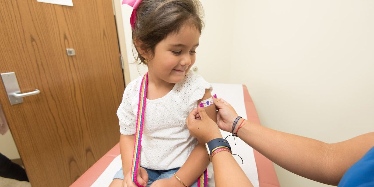 Young girl in a modern clinic calmly receiving a colourful bandage on her upper arm after a vaccination, healthcare provider in blue scrubs applying the bandage
