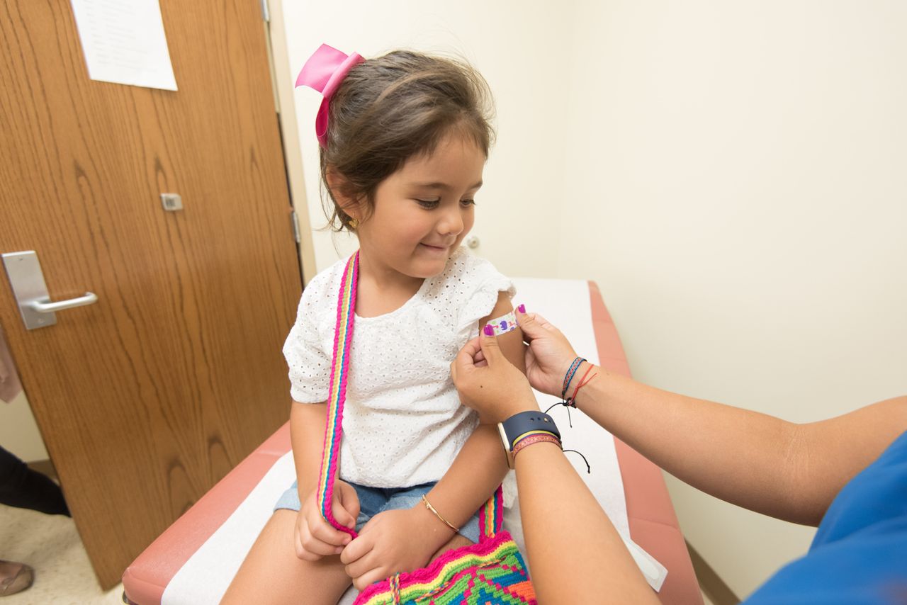 Young girl in a modern clinic calmly receiving a colourful bandage on her upper arm after a vaccination, healthcare provider in blue scrubs applying the bandage