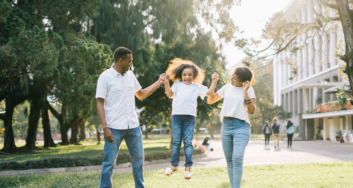 Happy well-to-do Indian parents swinging their young daughter in a park near a modern building