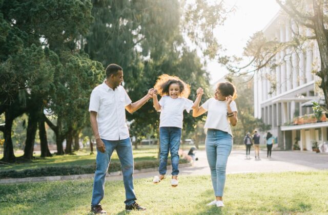 Happy well-to-do Indian parents swinging their young daughter in a park near a modern building
