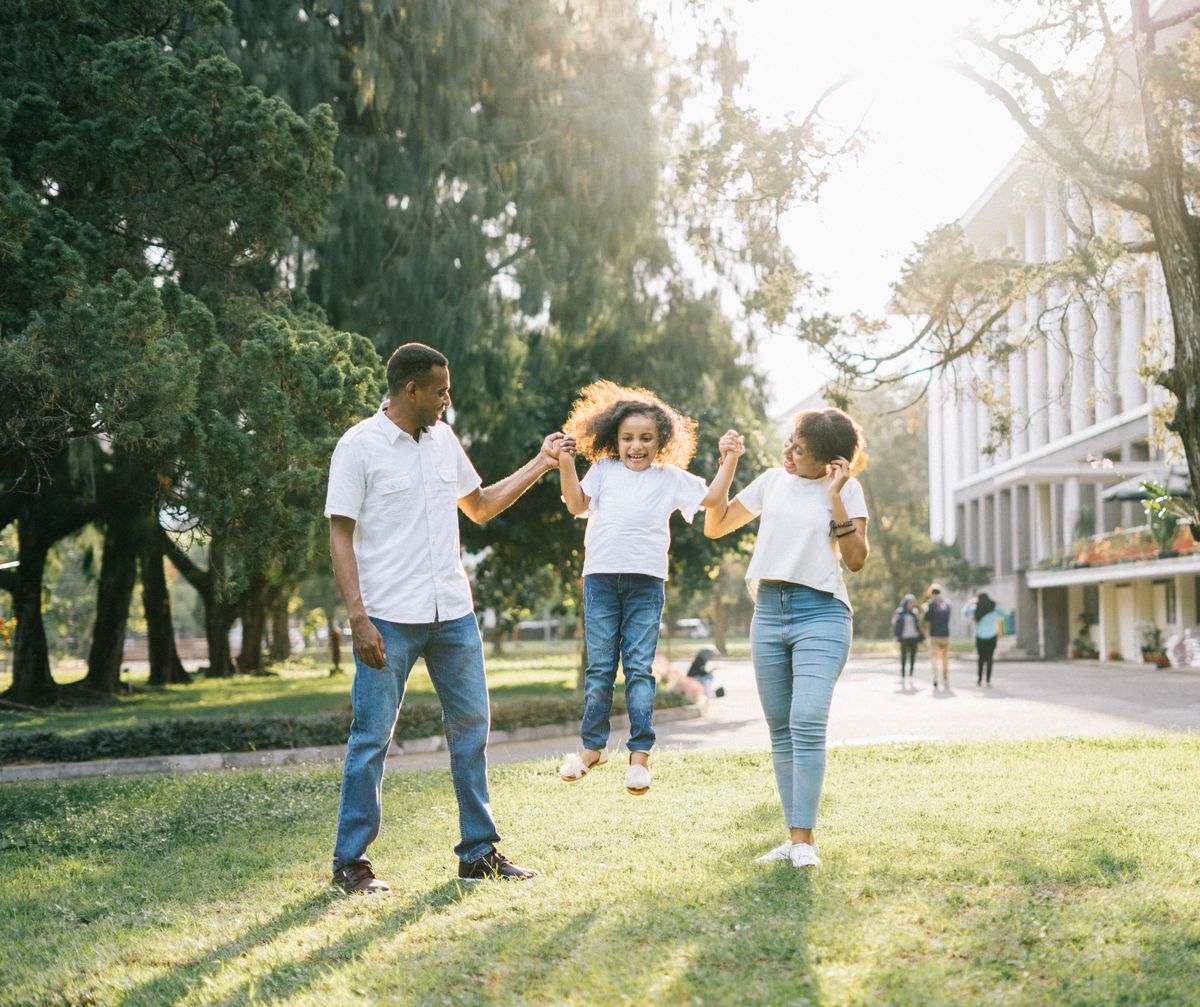 Happy well-to-do Indian parents swinging their young daughter in a park near a modern building