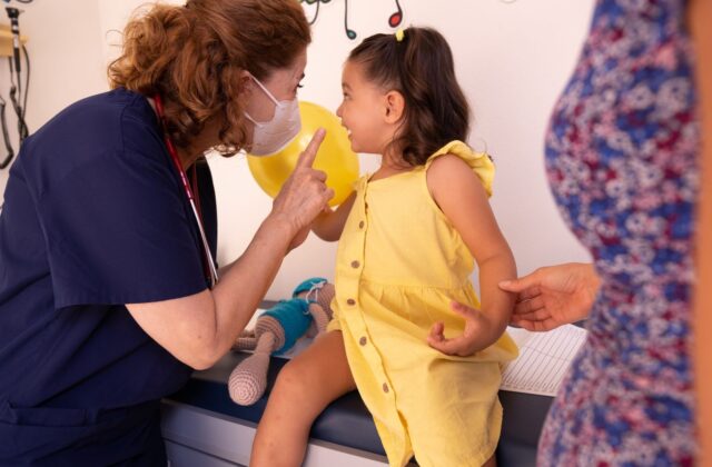 Well-dressed young girl in a yellow dress sitting cheerfully on a pediatric clinic exam table while a nurse in scrubs talks with her