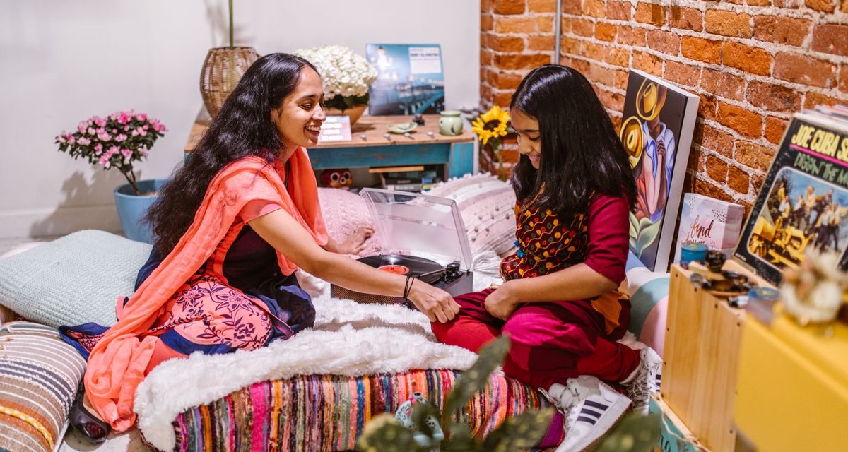 Well-dressed Indian mother and young daughter smiling together at home on colorful cushions beside a record player in a modern living room