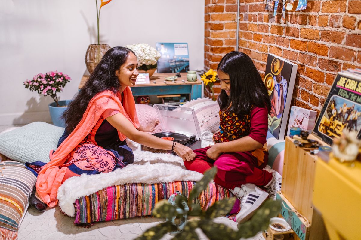Well-dressed Indian mother and young daughter smiling together at home on colorful cushions beside a record player in a modern living room