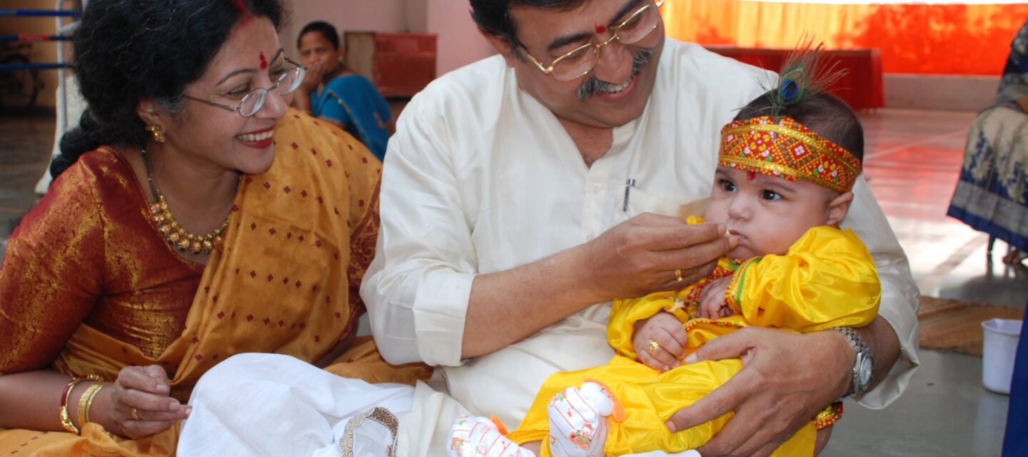 Smiling Indian family in festive silk saree and kurta gently feeding a baby in a yellow outfit a first taste of food at an annaprashan ceremony