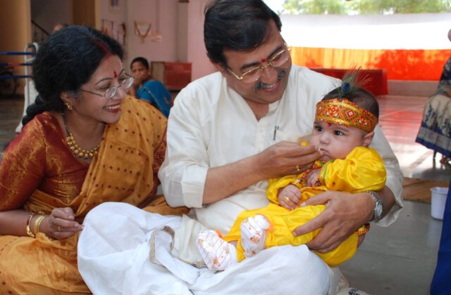 Smiling Indian family in festive silk saree and kurta gently feeding a baby in a yellow outfit a first taste of food at an annaprashan ceremony