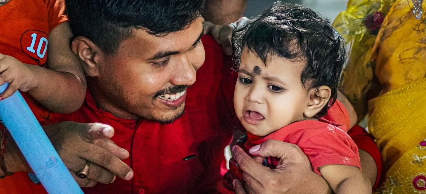 Smiling Indian father in a red shirt holding and comforting his crying toddler, who wears a red top and a traditional black dot on the forehead, at a family gathering
