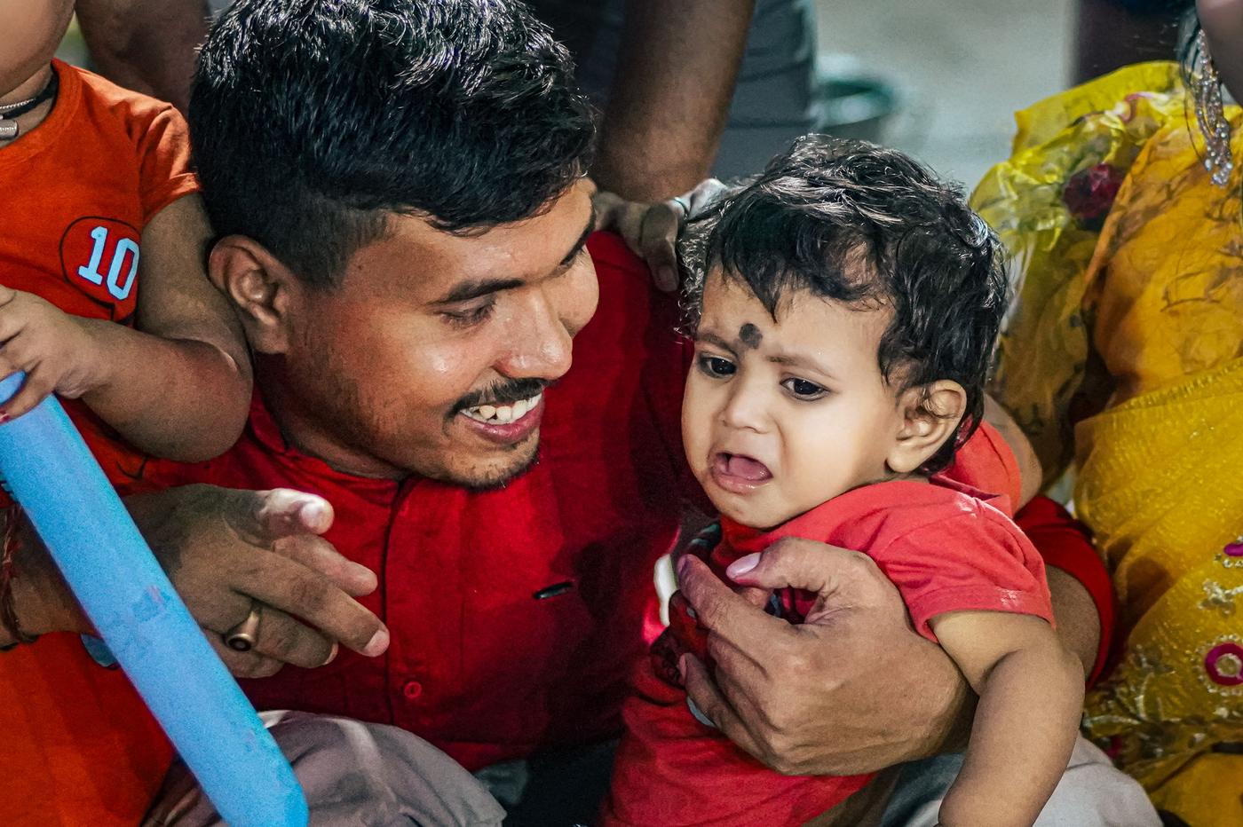 Smiling Indian father in a red shirt holding and comforting his crying toddler, who wears a red top and a traditional black dot on the forehead, at a family gathering