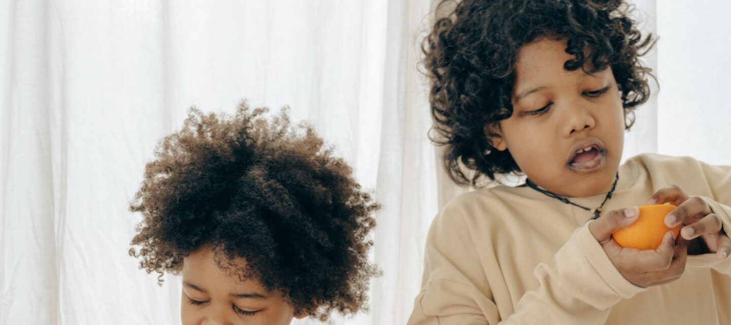 Two well-dressed children in a bright modern home peeling and eating fresh oranges by a sunlit curtain
