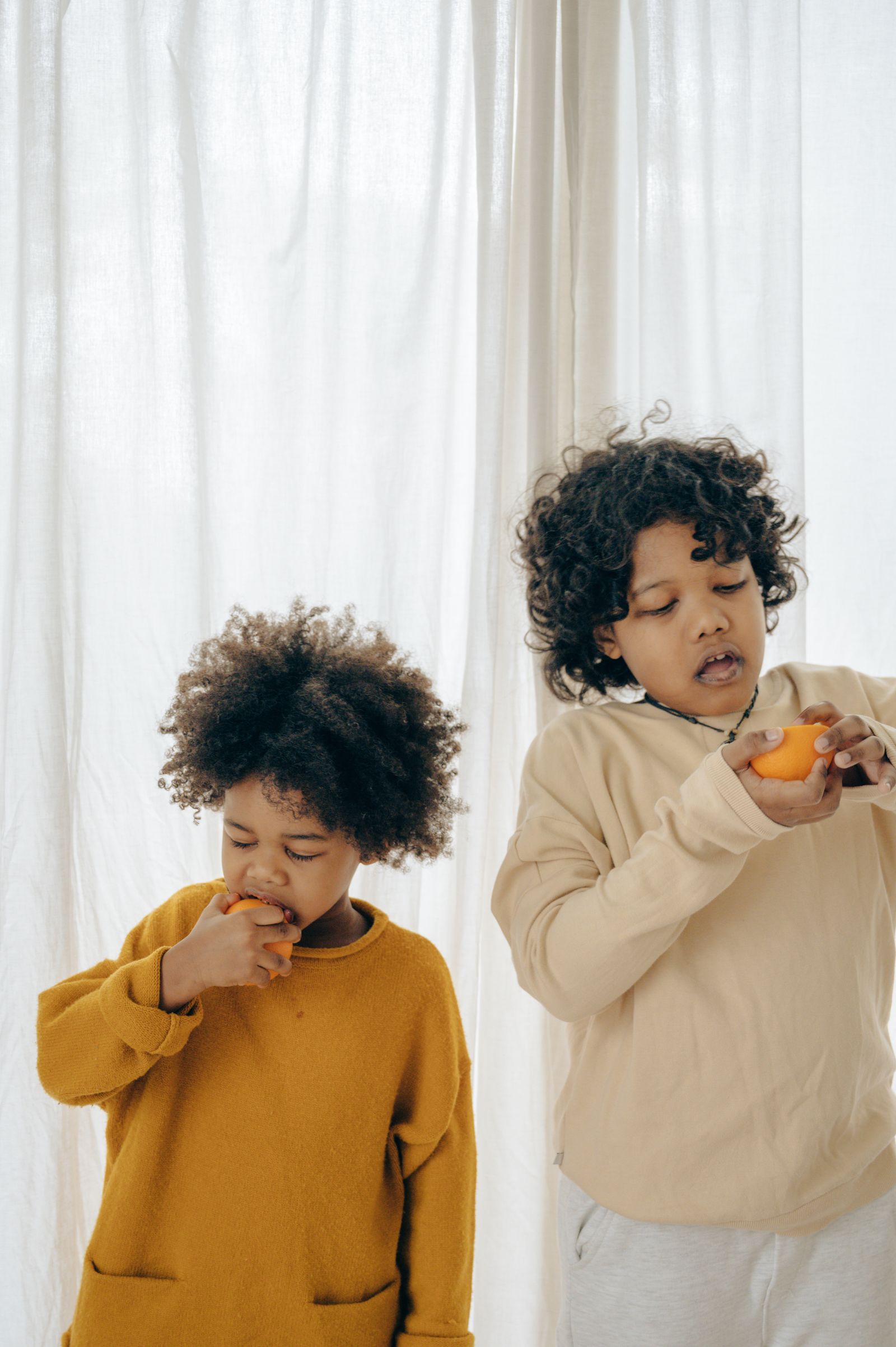 Two well-dressed children in a bright modern home peeling and eating fresh oranges by a sunlit curtain