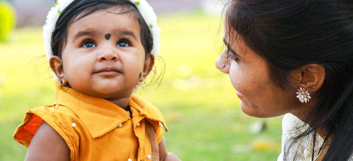 Indian mother smiling at her baby daughter outdoors in bright daylight, baby in a mustard dress with a soft headband—natural sunlight supporting vitamin D