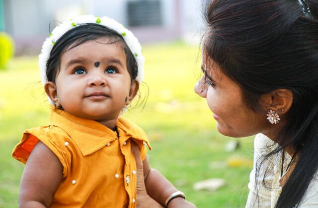 Indian mother smiling at her baby daughter outdoors in bright daylight, baby in a mustard dress with a soft headband—natural sunlight supporting vitamin D