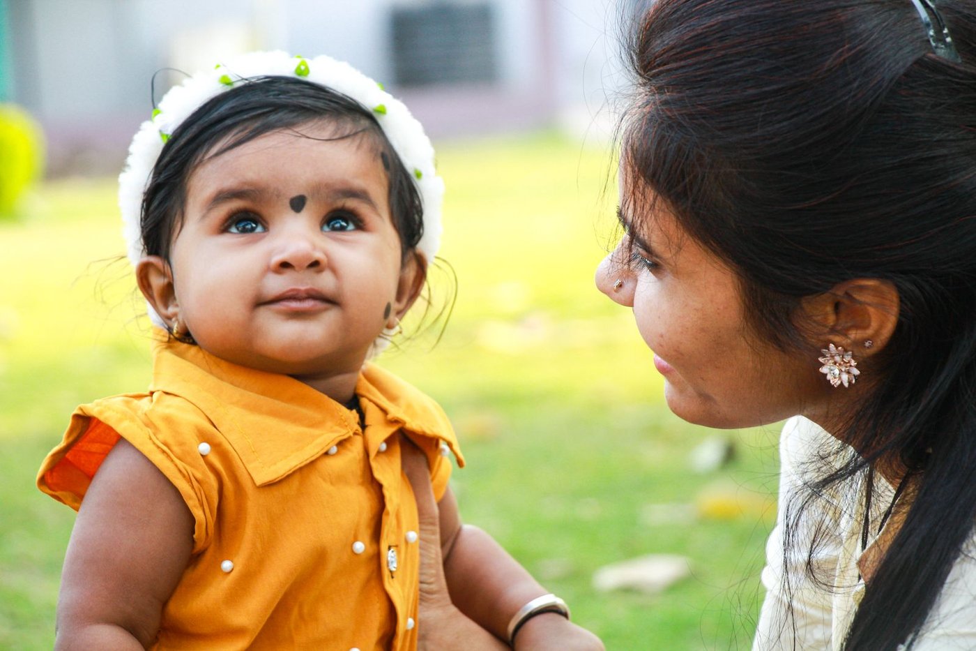Indian mother smiling at her baby daughter outdoors in bright daylight, baby in a mustard dress with a soft headband—natural sunlight supporting vitamin D