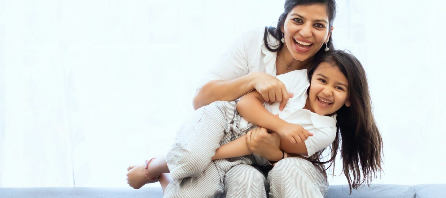 Smiling Indian mother hugging her laughing daughter on the sofa at home, a reminder that comfort and close care help when managing urinary tract infection in children
