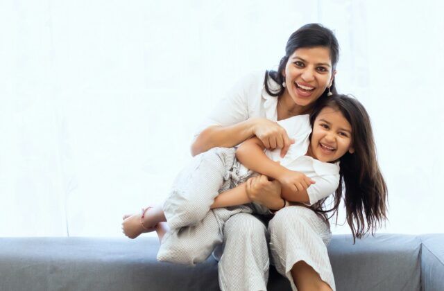 Smiling Indian mother hugging her laughing daughter on the sofa at home, a reminder that comfort and close care help when managing urinary tract infection in children