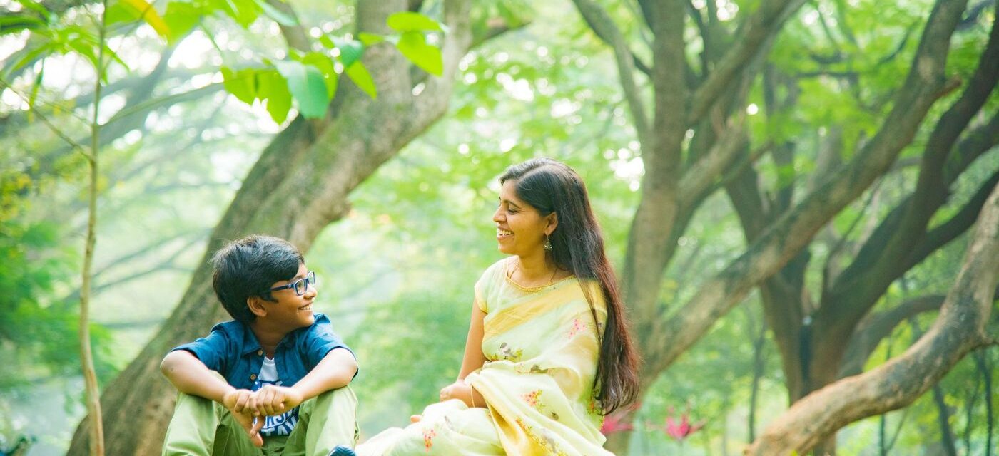 Smiling Indian mother in a pale yellow saree sitting on the grass in a green park with her son in glasses, blue shirt and sneakers, after outdoor play