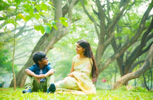 Smiling Indian mother in a pale yellow saree sitting on the grass in a green park with her son in glasses, blue shirt and sneakers, after outdoor play