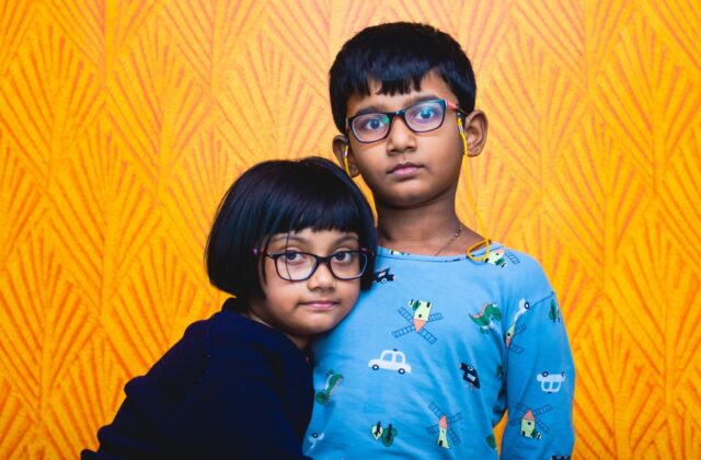 Indian brother and sister, both wearing eyeglasses, standing close together against a bright orange patterned wall