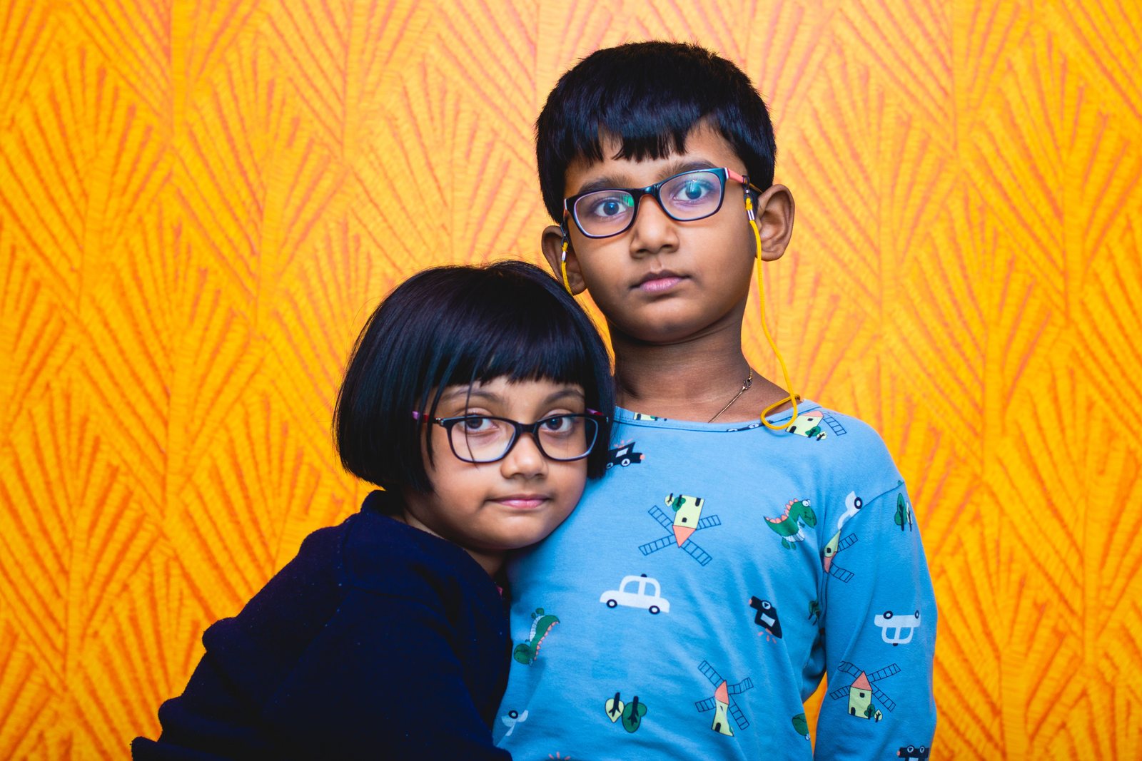 Indian brother and sister, both wearing eyeglasses, standing close together against a bright orange patterned wall