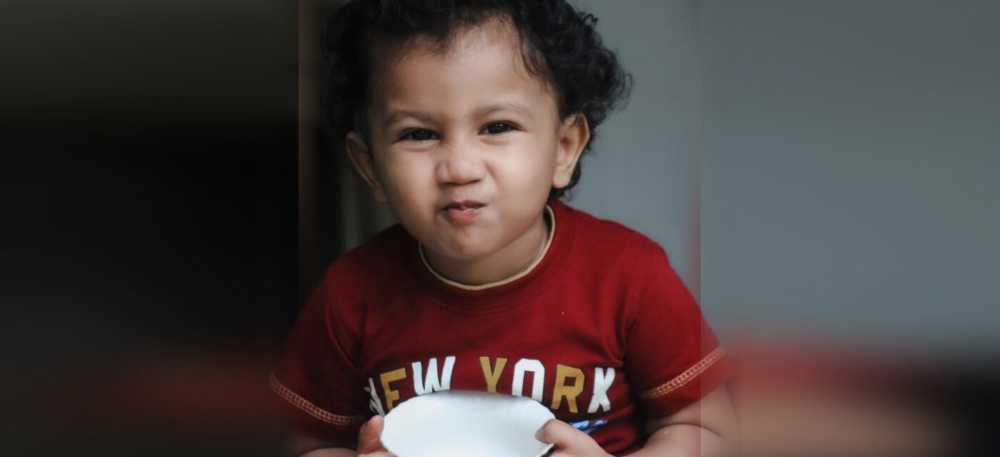 Indian toddler with curly hair in a red T-shirt holding a white bowl and pressing his lips together with a playful, fussy expression at mealtime