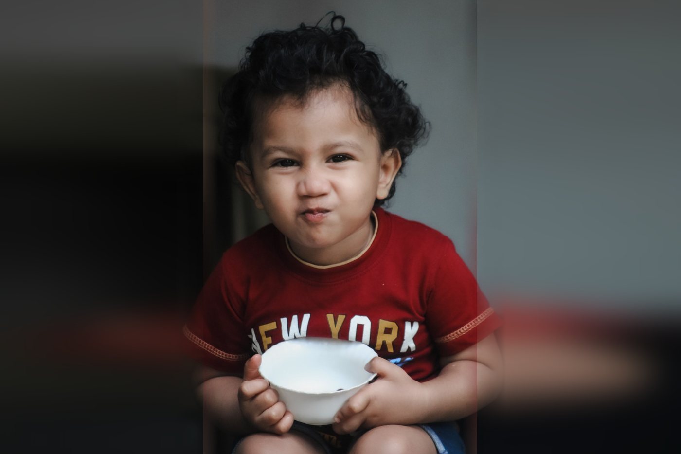 Indian toddler with curly hair in a red T-shirt holding a white bowl and pressing his lips together with a playful, fussy expression at mealtime