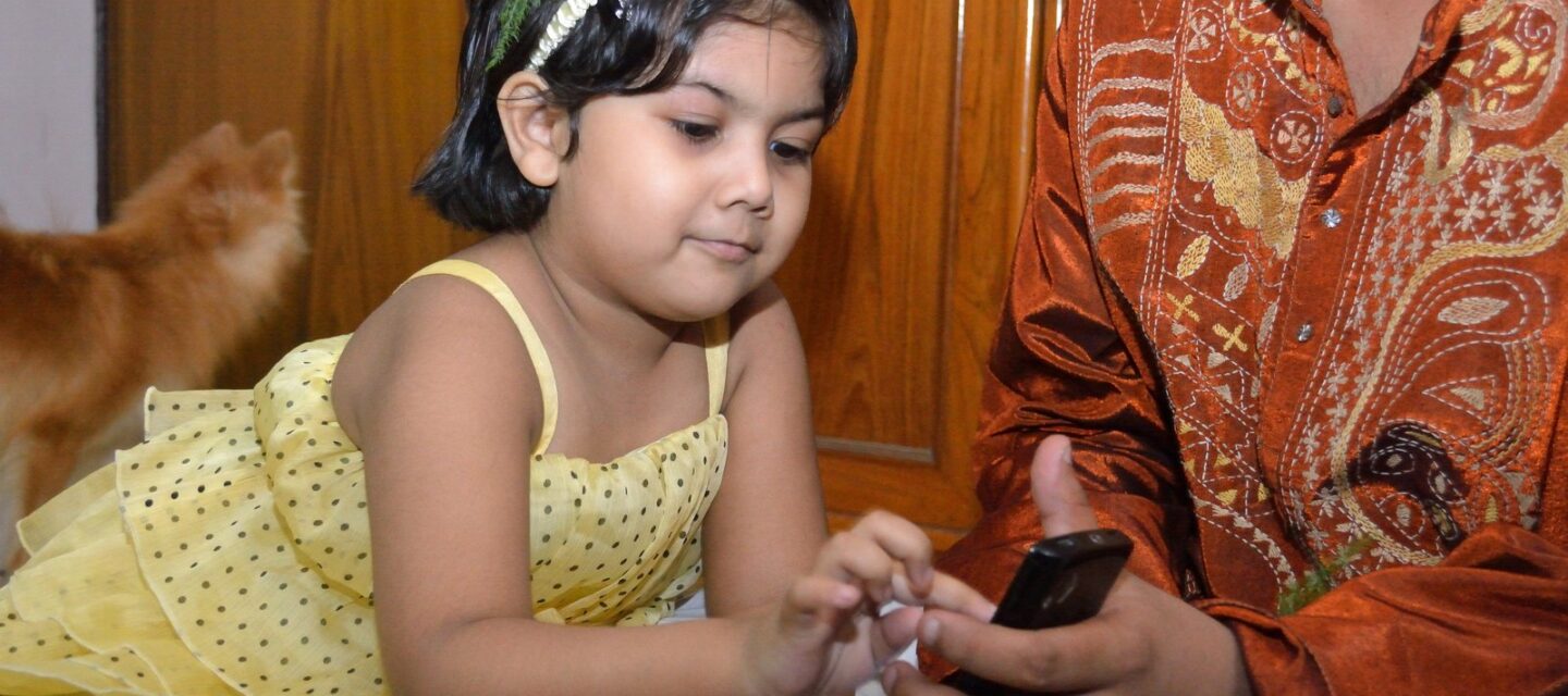 Indian toddler girl in a yellow party frock with a red rose in her hair touching a smartphone held by a family member in an embroidered silk kurta at home