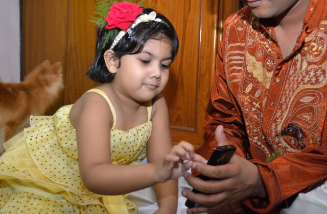 Indian toddler girl in a yellow party frock with a red rose in her hair touching a smartphone held by a family member in an embroidered silk kurta at home