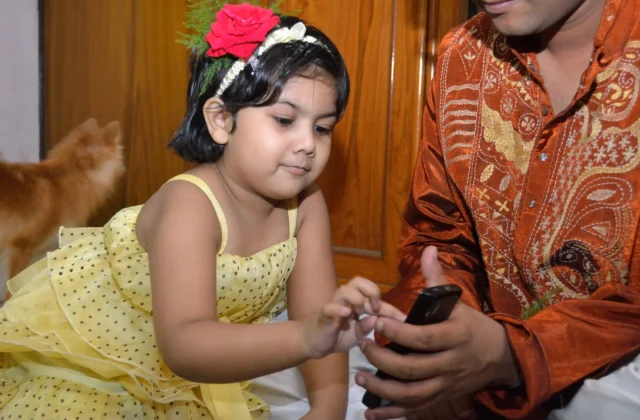 Indian toddler girl in a yellow party frock with a red rose in her hair touching a smartphone held by a family member in an embroidered silk kurta at home
